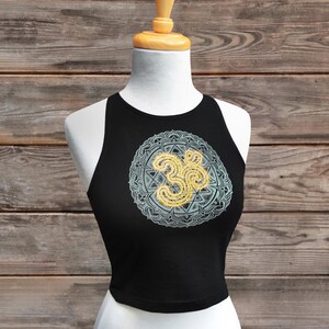 May include: Black crop top with a round Om symbol design in yellow and light blue. The top is displayed on a white mannequin against a wooden background. The Om symbol is a spiritual icon in Hinduism.