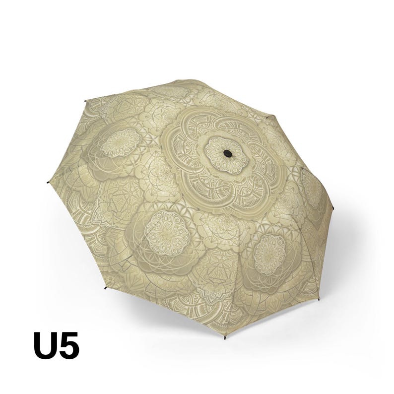 May include: A collapsible umbrella featuring a light beige canopy with a repeating intricate mandala pattern in darker shades of beige and cream. The center of the canopy has a small black circular hub. The text U5 is visible in the bottom left.
