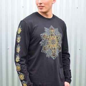 May include: Black long-sleeve shirt with a geometric design in gold and gray on the front. The sleeves have a repeating pattern of gold and gray geometric shapes.