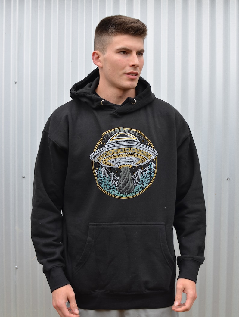 May include: Black hooded sweatshirt with a large, circular graphic of a UFO hovering over a mountain landscape. The UFO is detailed with gold and white accents, and the mountains are depicted in shades of blue and green.