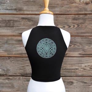 May include: Black crop top with a light blue geometric design on the back. The top is sleeveless and has a high neckline. The design is a detailed mandala pattern.