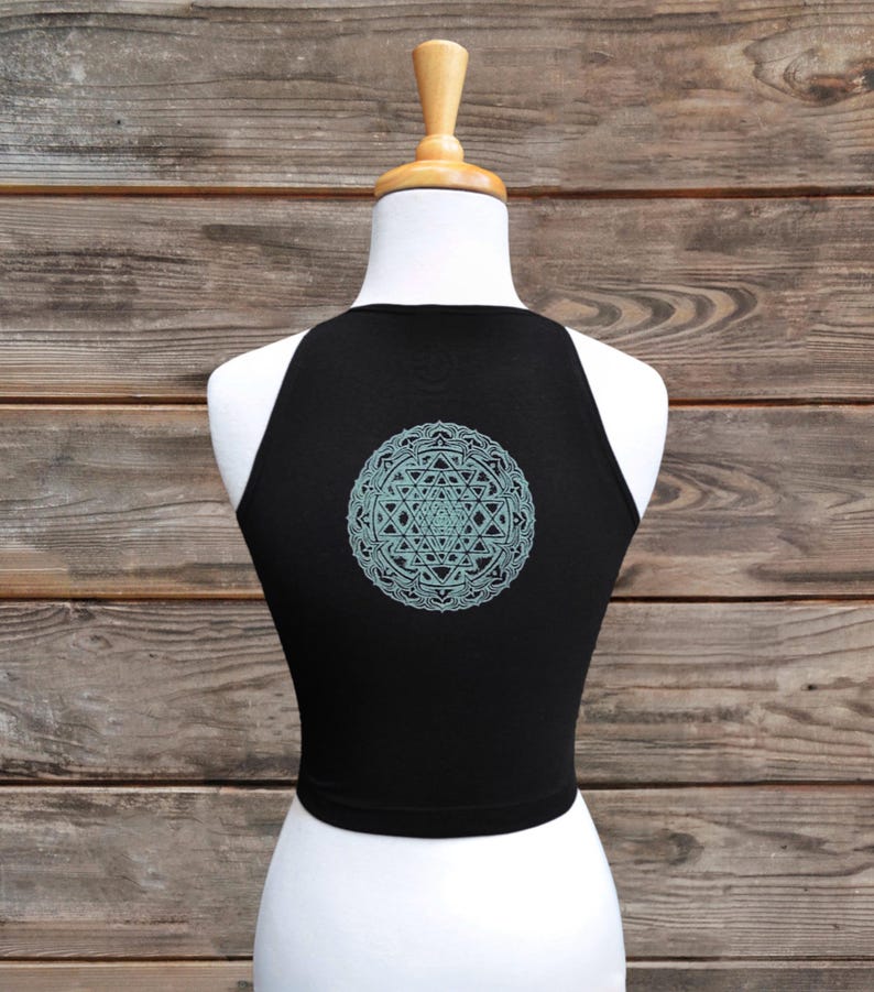 May include: Black crop top with a light blue geometric design on the back. The top is sleeveless and has a high neckline. The design is a detailed mandala pattern.