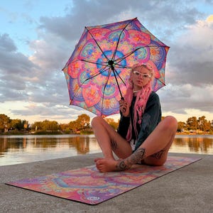 May include: A vibrant umbrella and a matching rectangular yoga mat, both featuring a complex, symmetrical mandala pattern in shades of bright pink, purple, orange, and blue. The umbrella has a black frame and handle, while the mat lies flat.