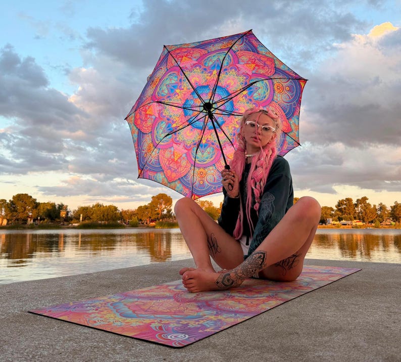May include: A vibrant umbrella and a matching rectangular yoga mat, both featuring a complex, symmetrical mandala pattern in shades of bright pink, purple, orange, and blue. The umbrella has a black frame and handle, while the mat lies flat.