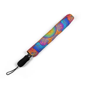 May include: A compact, closed umbrella featuring a vibrant, multi-colored geometric pattern in shades of blue, orange, pink, and yellow. The handle is black and cylindrical, equipped with a thin black wrist strap for carrying and secure storage.