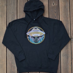 May include: Black hooded sweatshirt with a front pocket and drawstring. The sweatshirt features a colorful embroidered design of a UFO hovering over a mountain landscape. The UFO is detailed with yellow, white, and blue accents.
