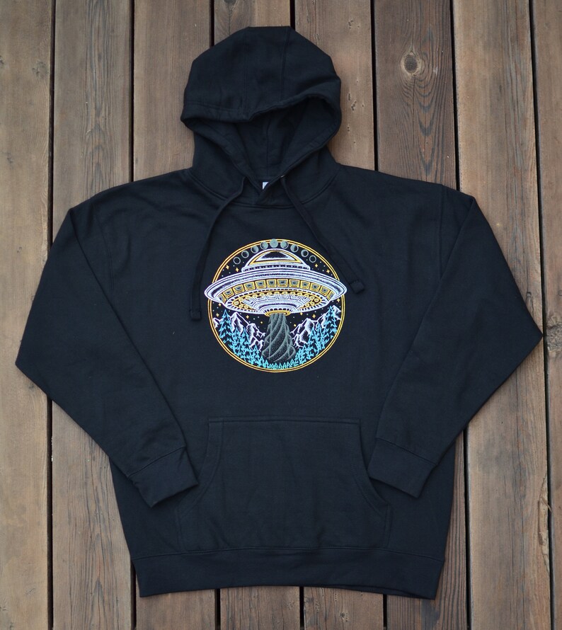 May include: Black hooded sweatshirt with a front pocket and drawstring. The sweatshirt features a colorful embroidered design of a UFO hovering over a mountain landscape. The UFO is detailed with yellow, white, and blue accents.