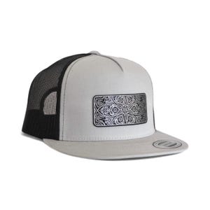 May include: A grey and black trucker hat with a silver decorative patch. The hat has a white front panel, a black mesh back, and a curved brim. The patch features an intricate, symmetrical design.