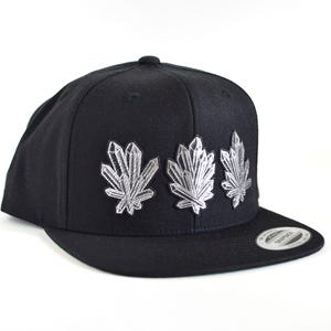 Może przedstawiać: Czarna czapka typu snapback z trzema wyhaftowanymi srebrnymi wzorami skupisk kryształów na przednim panelu. Czapka posiada usztywnianą koronę, płaski daszek oraz okrągłą naklejkę na daszku z napisem Snapback i Yupoong Classics.