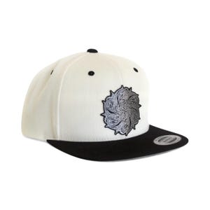 Può includere: Un cappellino snapback bianco con visiera nera e un bottone nero sulla parte superiore. Il pannello frontale presenta una toppa circolare con un motivo a vortice argentato dettagliato e complesso. Un piccolo adesivo è visibile sulla visiera.