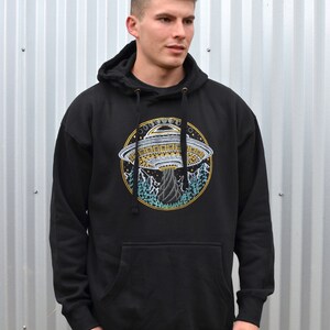 May include: Black hooded sweatshirt with a front pocket and drawstring. The sweatshirt features a circular embroidered design of a UFO, mountains, and trees. The design is in shades of yellow, blue, and white.