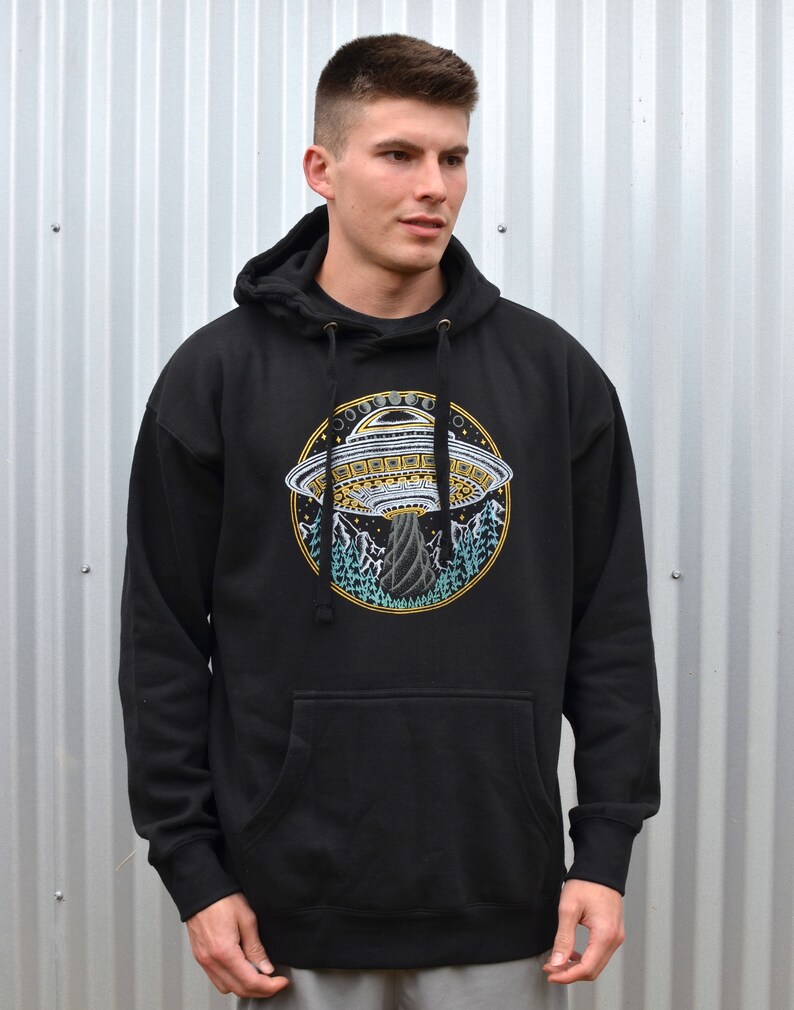 May include: Black hooded sweatshirt with a front pocket and drawstring. The sweatshirt features a circular embroidered design of a UFO, mountains, and trees. The design is in shades of yellow, blue, and white.