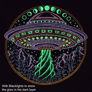 May include: Black graphic design featuring a purple UFO emitting a green beam over a mountain range and trees. The design includes a gold border and glowing green accents. Text at the bottom reads, "With Blacklights to show the glow in the dark layer."