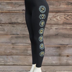 May include: Black leggings with seven chakra symbols in teal and gold down the side. The leggings are displayed on a mannequin leg against a wooden background. The symbols are in a vertical line.