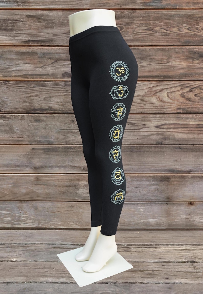 May include: Black leggings with seven chakra symbols in teal and gold down the side. The leggings are displayed on a mannequin leg against a wooden background. The symbols are in a vertical line.