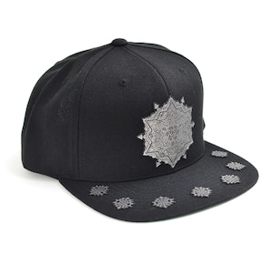 May include: Black snapback hat with a silver, ornate starburst design on the front and smaller starbursts on the brim. The hat is made of a textured fabric and has a flat brim.