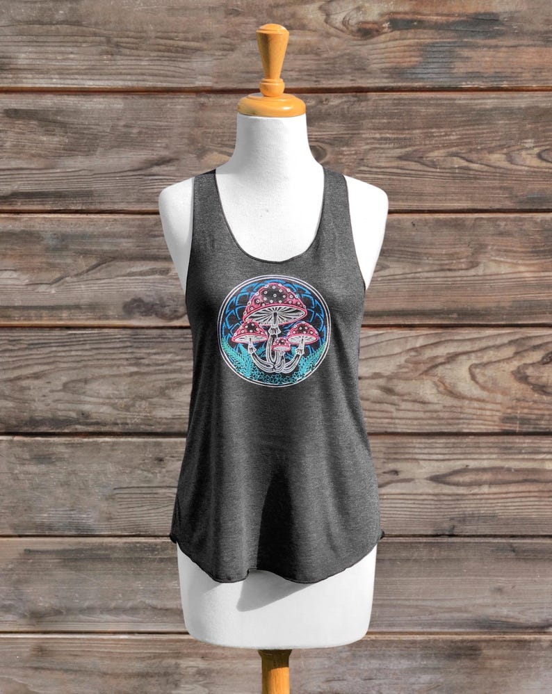 May include: Dark grey tank top with a circular graphic of mushrooms in pink, blue, and white. The top is displayed on a mannequin. The background is a wooden panel. The tank top is a casual style.