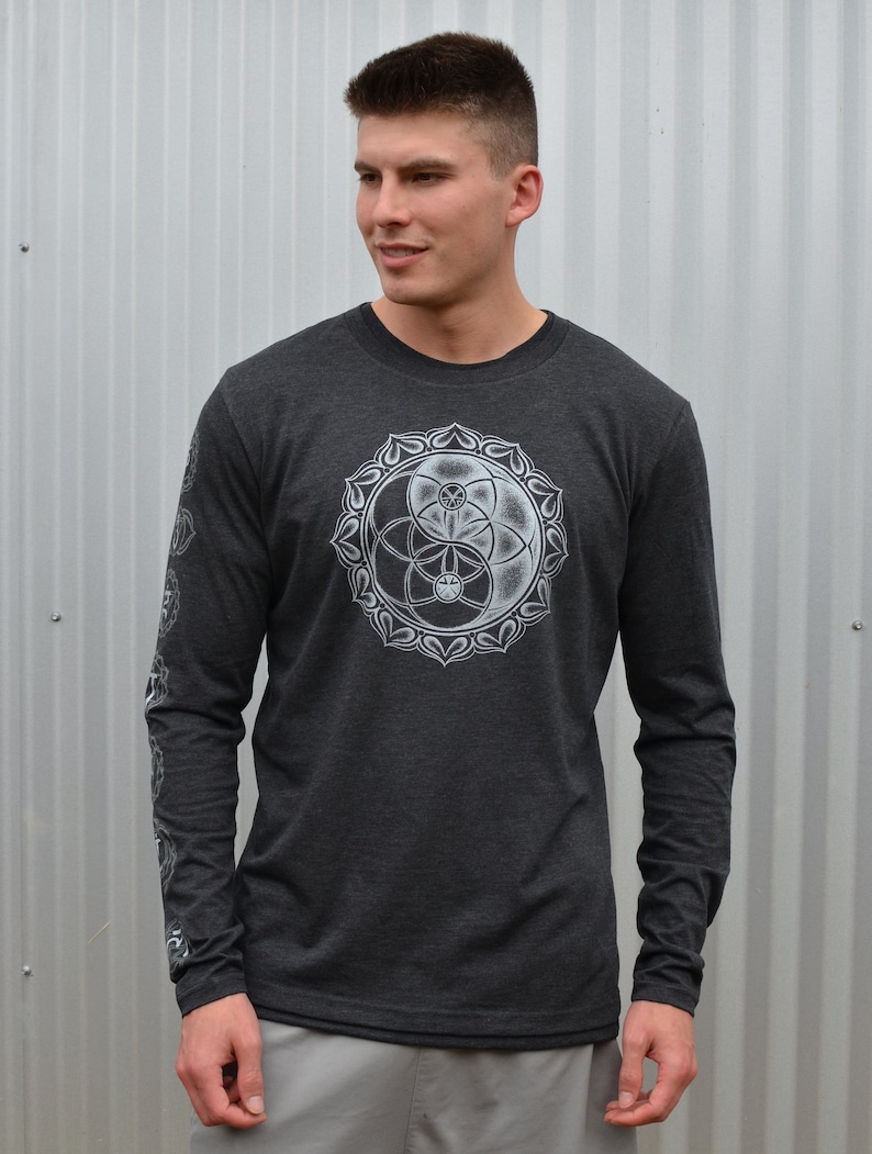 May include: A dark heather grey long-sleeve shirt with a white graphic of a yin yang symbol with a flower of life design inside. The shirt has a crew neck.