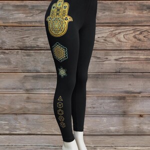 May include: Black leggings with gold and teal geometric designs. The leggings feature a Hamsa hand, a flower of life, and other geometric shapes. The leggings are displayed on a mannequin.