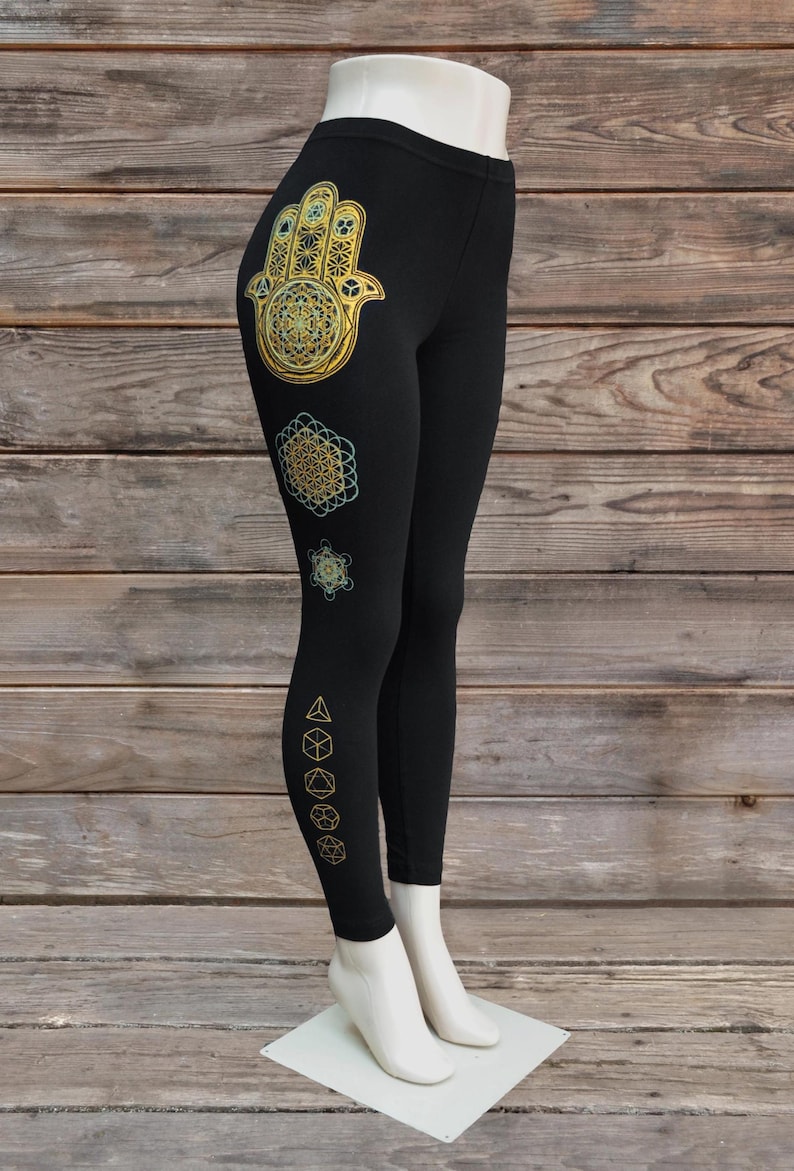 May include: Black leggings with gold and teal geometric designs. The leggings feature a Hamsa hand, a flower of life, and other geometric shapes. The leggings are displayed on a mannequin.