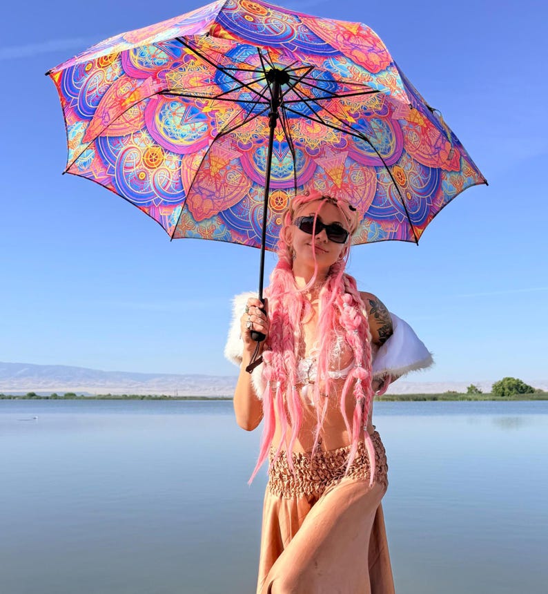 May include: A large, open umbrella featuring a vibrant, multi-colored mandala pattern in shades of pink, purple, blue, and orange. The umbrella has a black central shaft and black metal ribs supporting the canopy, which displays intricate geometric designs.