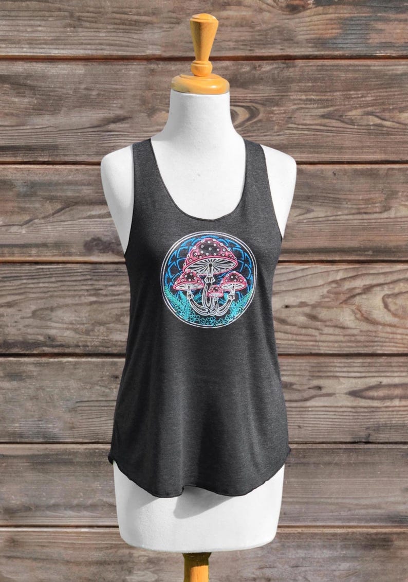 May include: Dark grey vest top featuring a circular design with pink and white mushrooms set against a blue and green background. The top is displayed on a mannequin against a wooden backdrop. The vest is made of a soft material.
