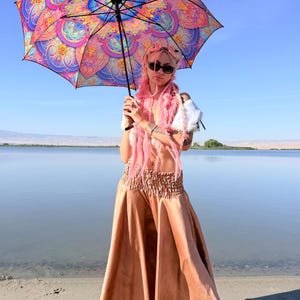 May include: A person with long pink hair and sunglasses holding a large umbrella featuring a vibrant, multi-colored mandala pattern in shades of pink, orange, blue, and purple. The person wears a white top and a long, flowing, metallic bronze skirt.