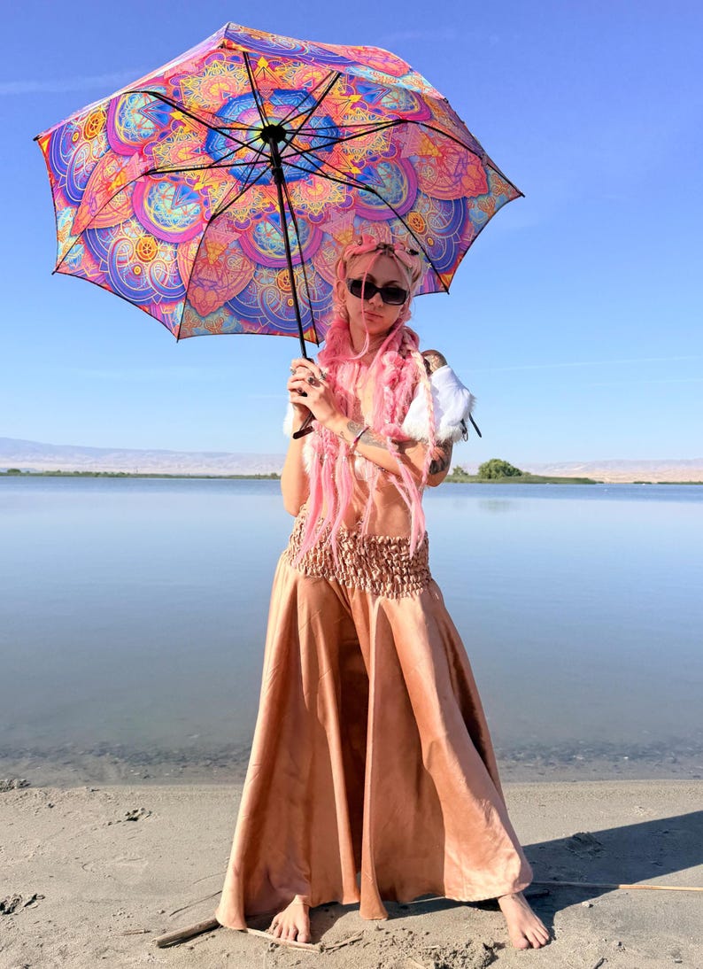 May include: A person with long pink hair and sunglasses holding a large umbrella featuring a vibrant, multi-colored mandala pattern in shades of pink, orange, blue, and purple. The person wears a white top and a long, flowing, metallic bronze skirt.