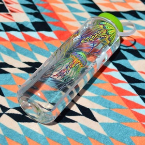 K&ouml;nnte beinhalten: Eine durchsichtige Plastikwasserflasche mit gr&uuml;nem Deckel und einem bunten Quallen-Design. Die Flasche liegt auf einem bunten, gemusterten Stoff.