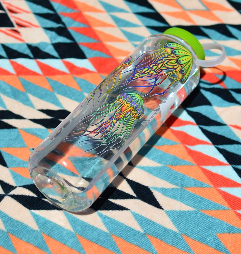 K&ouml;nnte beinhalten: Eine durchsichtige Plastikwasserflasche mit gr&uuml;nem Deckel und einem bunten Quallen-Design. Die Flasche liegt auf einem bunten, gemusterten Stoff.