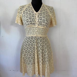 May include: A vintage cream-colored lace dress with a floral pattern. The dress has a V-neckline, short sleeves, and a fitted bodice. The skirt is full and flowing.