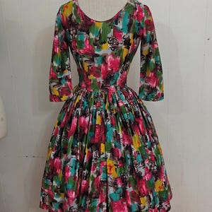 May include: A vintage 1950s style dress with a full skirt and 3/4 sleeves. The dress is made of a colorful cotton fabric with an abstract floral print in shades of pink, yellow, green, blue, and brown.