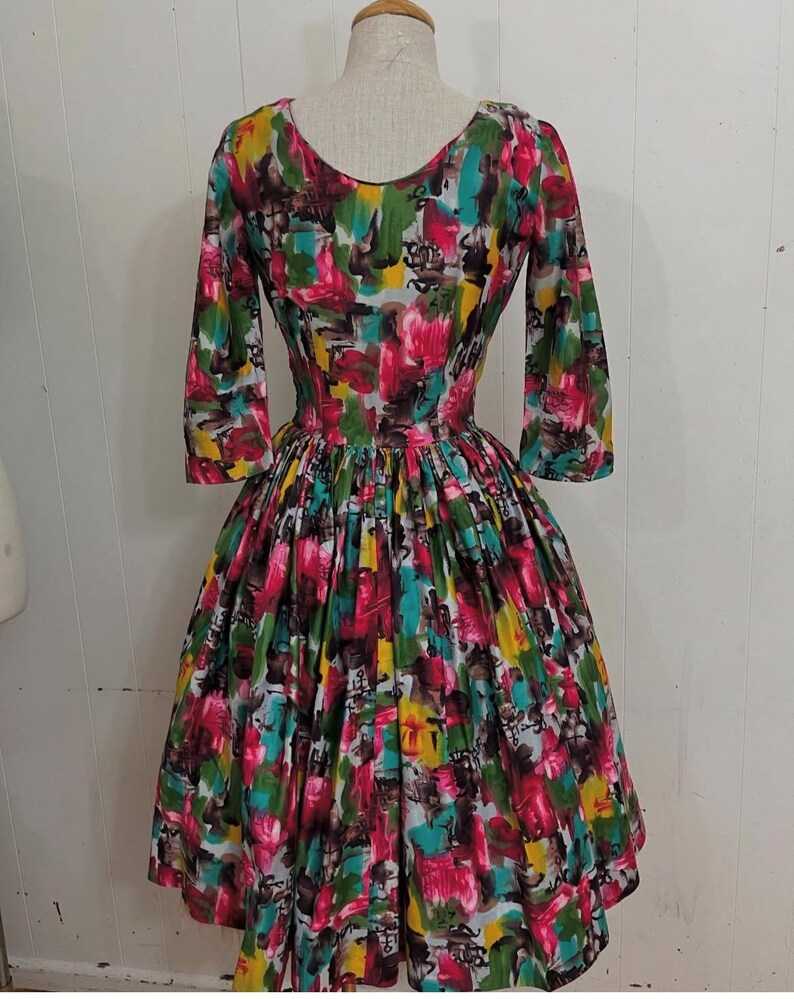 May include: A vintage 1950s style dress with a full skirt and 3/4 sleeves. The dress is made of a colorful cotton fabric with an abstract floral print in shades of pink, yellow, green, blue, and brown.