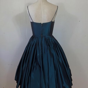 May include: A teal vintage dress with a fitted bodice and a full, flared skirt. The dress has thin spaghetti straps and a zip closure at the back. The fabric has a subtle sheen. The dress is displayed on a mannequin. The dress is approximately 100 cm in length.