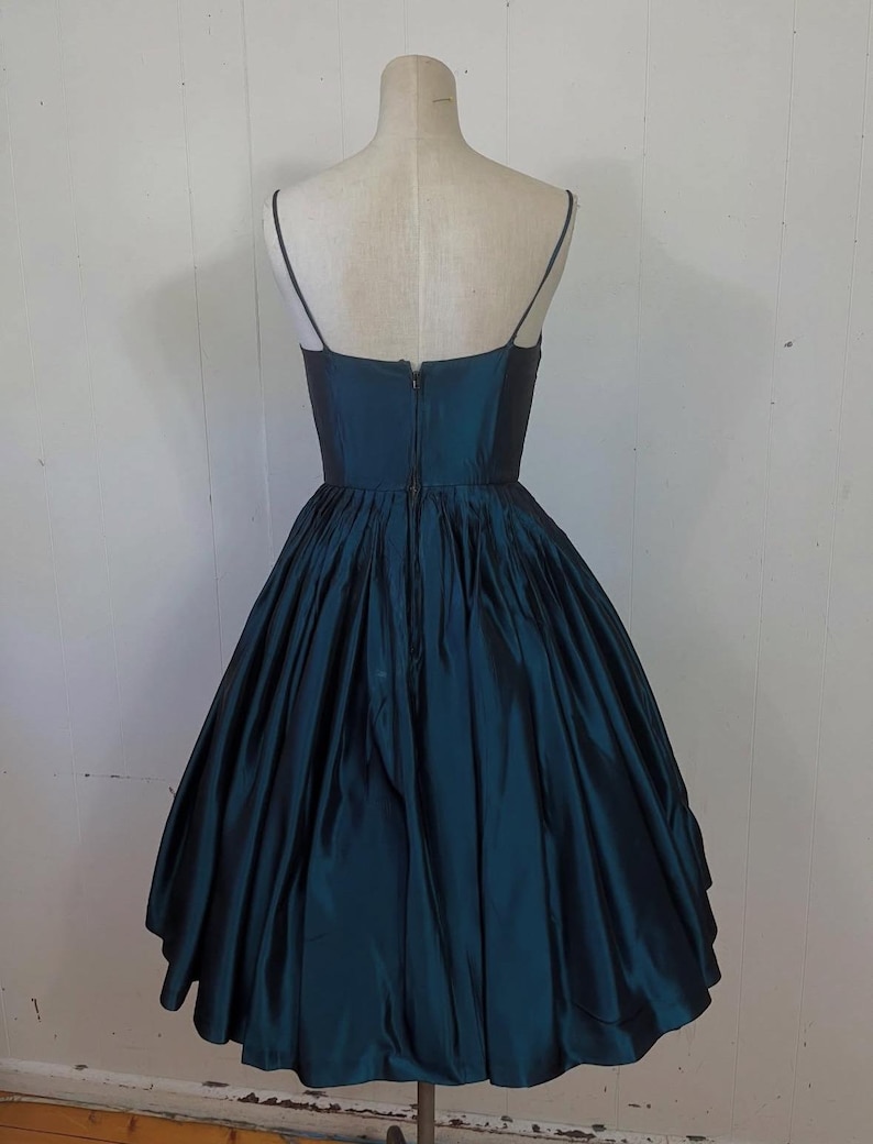 May include: A teal vintage dress with a fitted bodice and a full, flared skirt. The dress has thin spaghetti straps and a zip closure at the back. The fabric has a subtle sheen. The dress is displayed on a mannequin. The dress is approximately 100 cm in length.