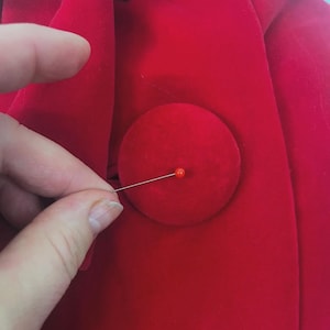 May include: Close-up of a bright red fabric with a large, round button. A straight pin with a red head is inserted into the button. The fabric appears to be a soft material, possibly velvet or a similar plush textile.