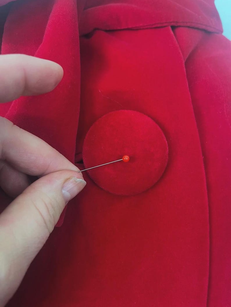 May include: Close-up of a bright red fabric with a large, round button. A straight pin with a red head is inserted into the button. The fabric appears to be a soft material, possibly velvet or a similar plush textile.