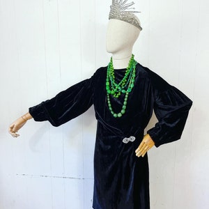May include: A black velvet dress with a wrap style and long sleeves. The dress features a silver brooch at the waist and is worn with a layered green beaded necklace. The model is wearing a silver crown.
