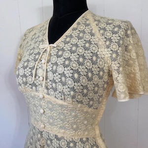 May include: A cream-colored lace dress with a V-neckline, short sleeves, and a button-up front. The dress has a delicate floral pattern and a fitted bodice.