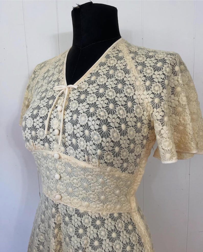 May include: A cream-colored lace dress with a V-neckline, short sleeves, and a button-up front. The dress has a delicate floral pattern and a fitted bodice.