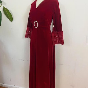 Può includere: Un lungo abito di velluto rosso con scollo a V e una cintura gioiello. L'abito ha maniche a 3/4 con bordature in pizzo.
