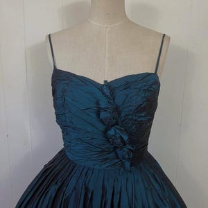 May include: A teal formal dress with a strapless bodice and a full skirt. The bodice has gathered fabric and a floral accent. The skirt is pleated, creating a voluminous silhouette. The dress is displayed on a mannequin.