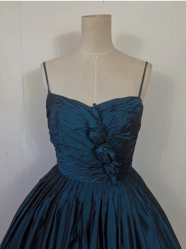 May include: A teal formal dress with a strapless bodice and a full skirt. The bodice has gathered fabric and a floral accent. The skirt is pleated, creating a voluminous silhouette. The dress is displayed on a mannequin.