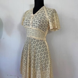 May include: A vintage cream-colored lace dress with a floral pattern. The dress has a V-neckline, short sleeves, and a fitted bodice. The skirt is full and flowing.