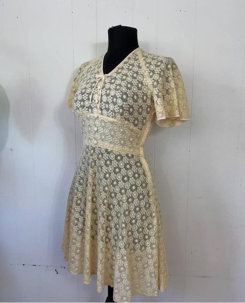 May include: A vintage cream-colored lace dress with a floral pattern. The dress has a V-neckline, short sleeves, and a fitted bodice. The skirt is full and flowing.