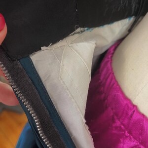 May include: Close-up of a garment's interior, showing a black fabric collar and a teal zipper. The lining is white with a quilted pattern, and a vibrant magenta fabric is visible. The zipper is silver.