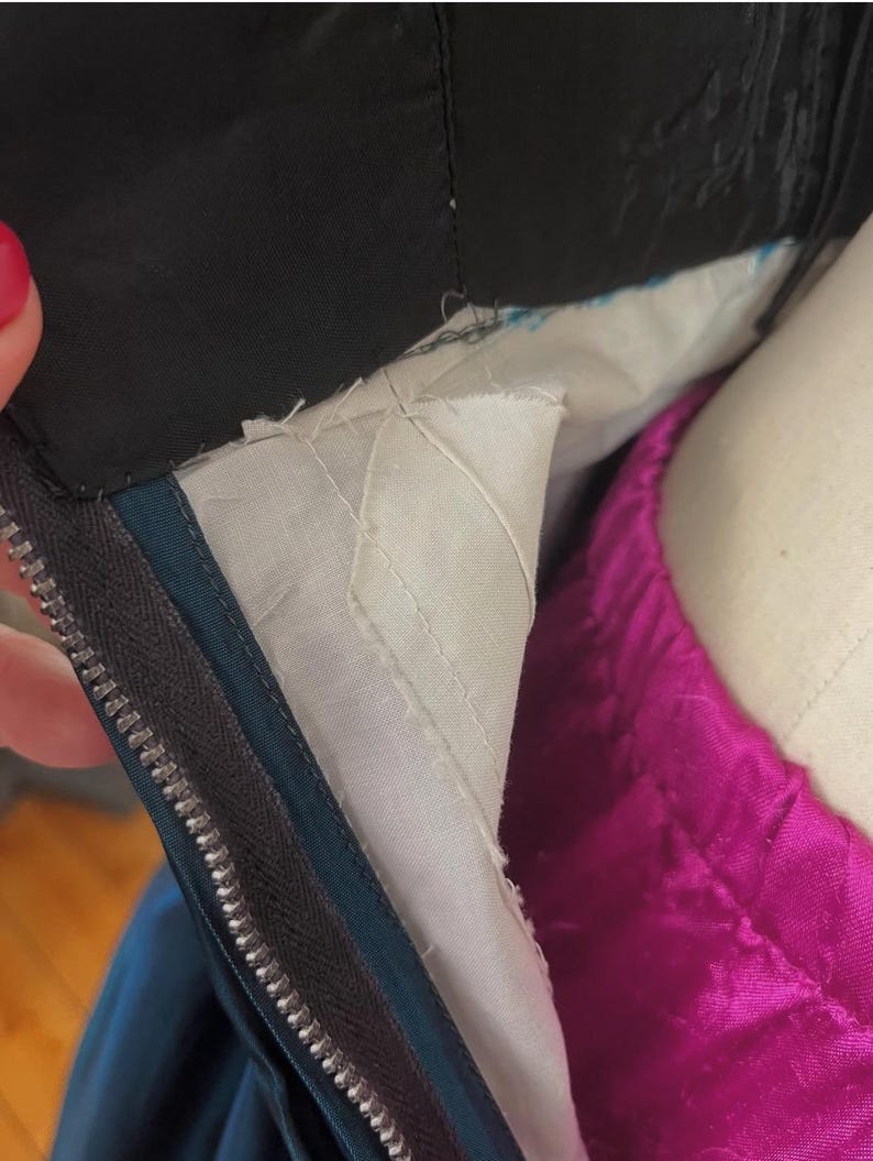 May include: Close-up of a garment's interior, showing a black fabric collar and a teal zipper. The lining is white with a quilted pattern, and a vibrant magenta fabric is visible. The zipper is silver.