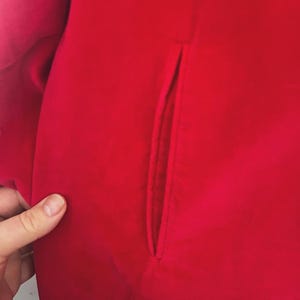 May include: Close-up of a vibrant red garment, possibly a coat or jacket, featuring a tailored pocket detail. The fabric appears smooth and solid, with a slight sheen. The image highlights the clean lines and rich color of the clothing item.