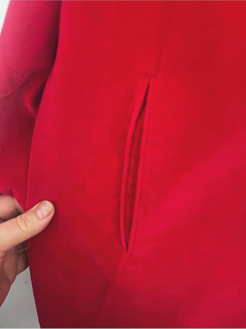 May include: Close-up of a vibrant red garment, possibly a coat or jacket, featuring a tailored pocket detail. The fabric appears smooth and solid, with a slight sheen. The image highlights the clean lines and rich color of the clothing item.
