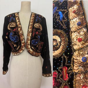 May include: Black velvet bolero jacket adorned with sequins and beads. The jacket has gold, red, and blue detailing, with a paisley pattern on the front. The edges are trimmed with gold sequins, giving it a vintage and ornate appearance.